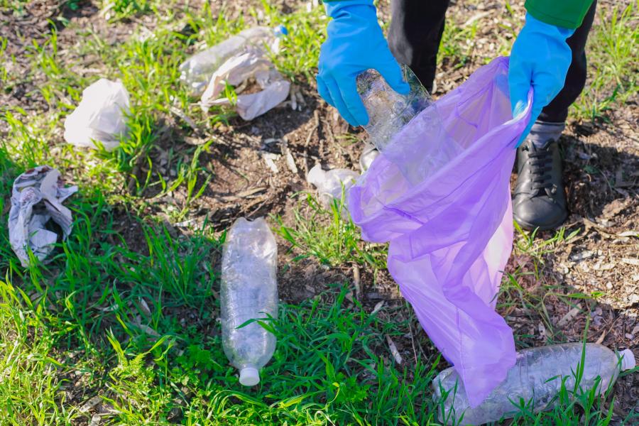 Close-up photo of a person with gloves on picking up plastic bottles and other litter from grass and putting it into a trash bag